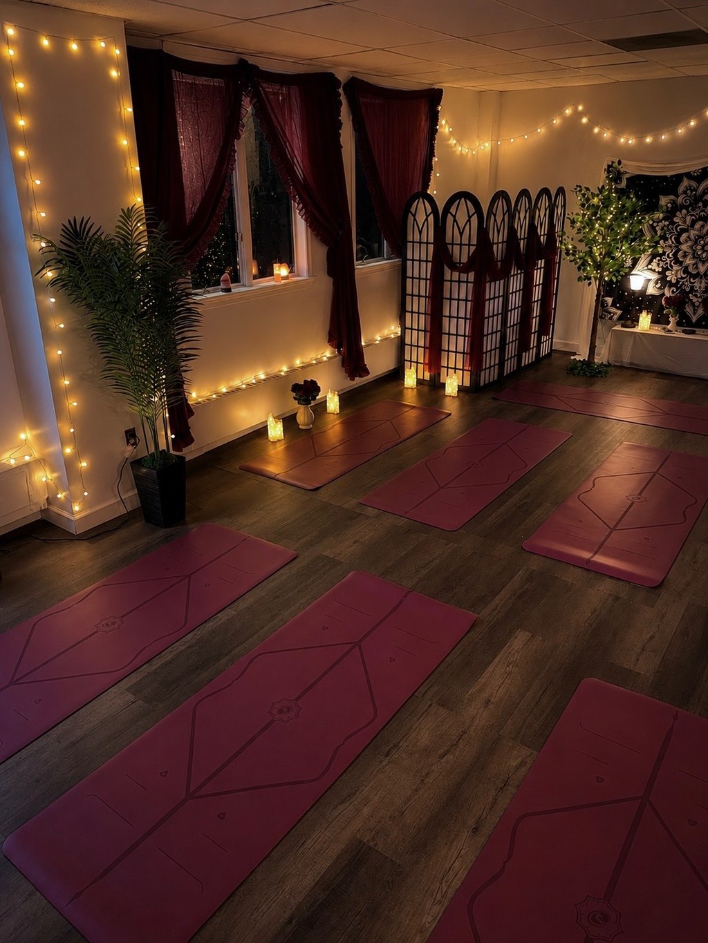 Interior of Sophia Selene Yoga studio at 17 West Jefferson Street, Suite 007, glowing at evening — burgundy yoga mats on a dark wood floor lit by candles, warm string lights along the walls, deep-red curtains, a folding screen with arched panels, and greenery in the corners.