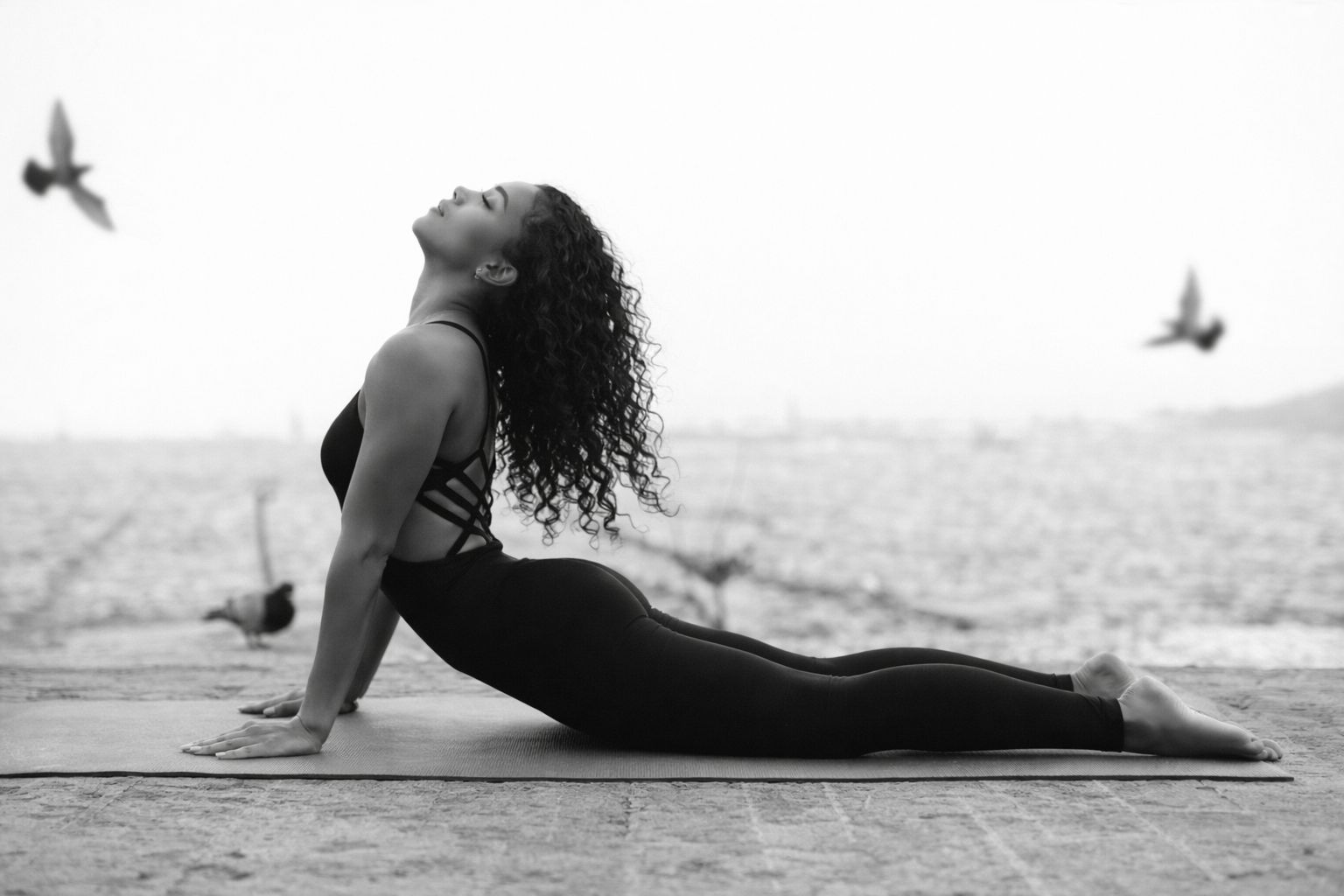 Sophia in upward-facing dog on the shore at dawn, birds in flight around her — black and white.