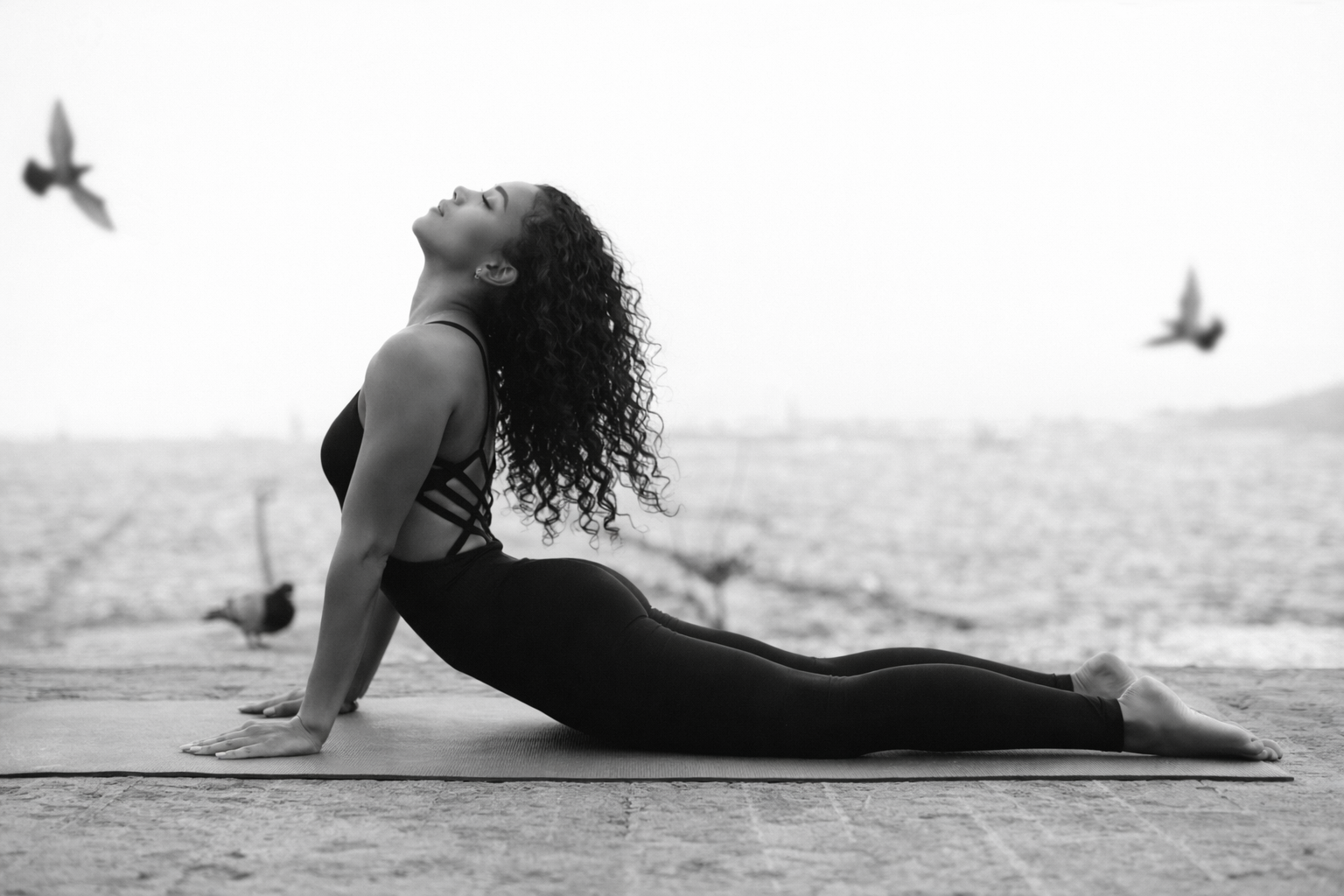 Sophia in upward-facing dog on the shore at dawn, birds in flight around her — black and white.