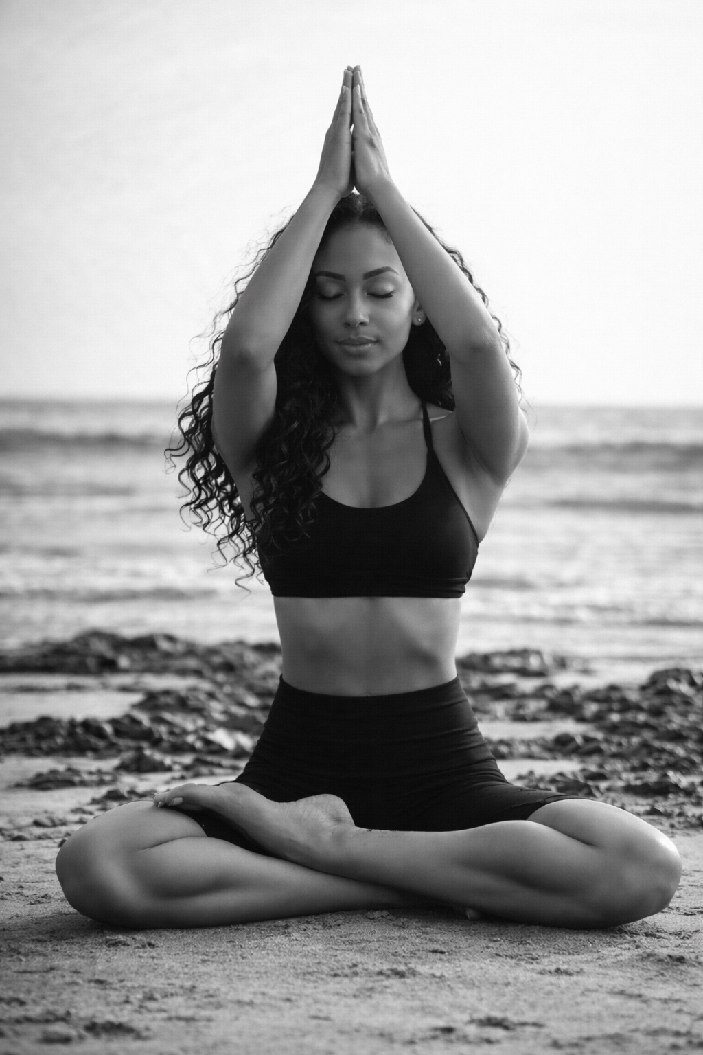 Sophia seated in lotus on the beach, palms pressed overhead in prayer — black and white.