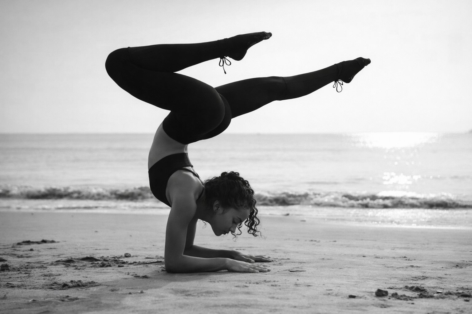 Sophia in a forearm scorpion balance on wet sand, ocean behind her — black and white.