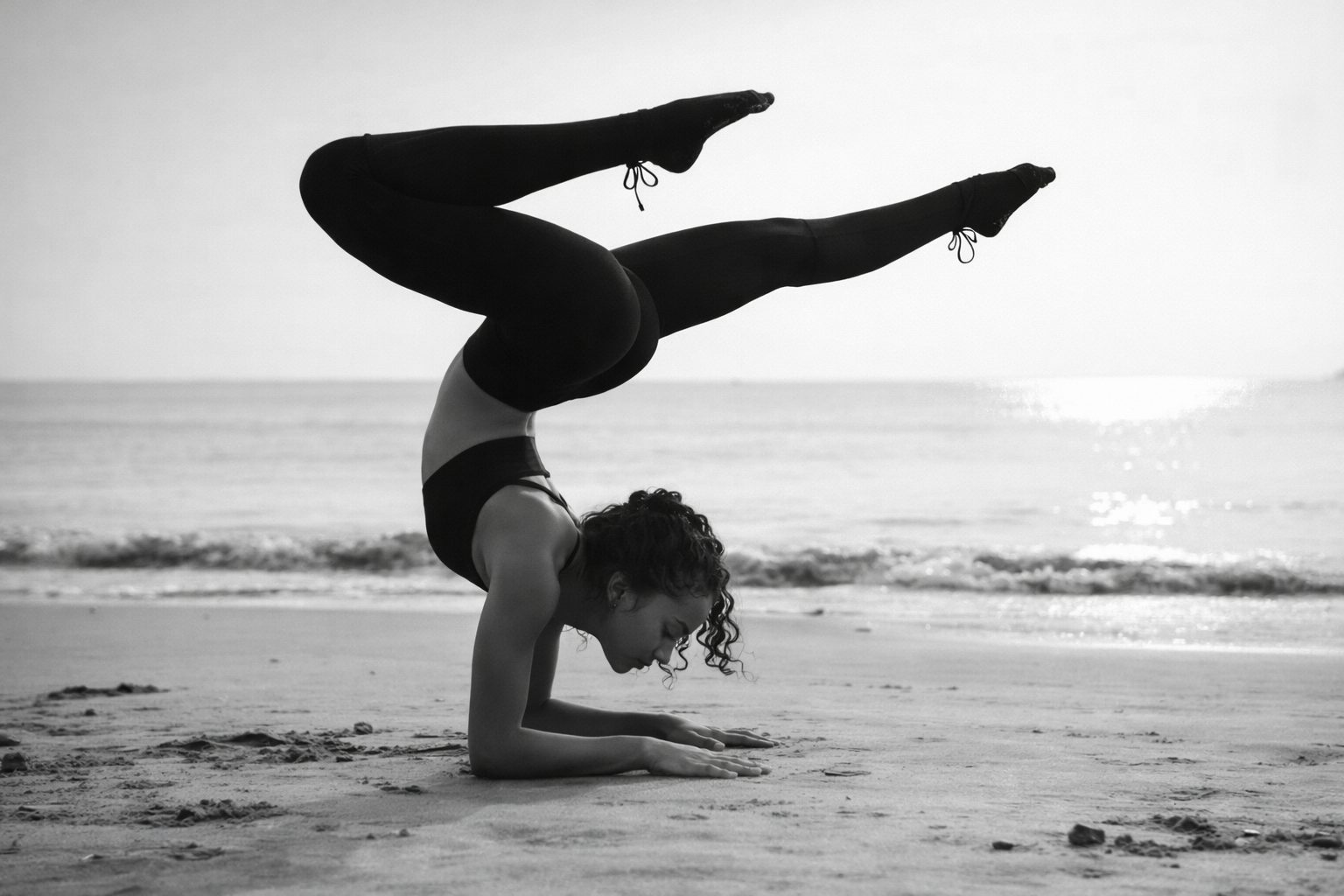 Sophia in a forearm scorpion balance on wet sand, ocean behind her — black and white.