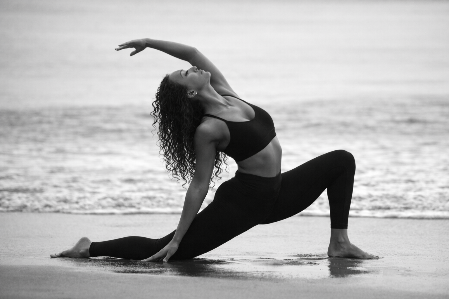 Sophia in a low lunge side bend at the water's edge — black and white.