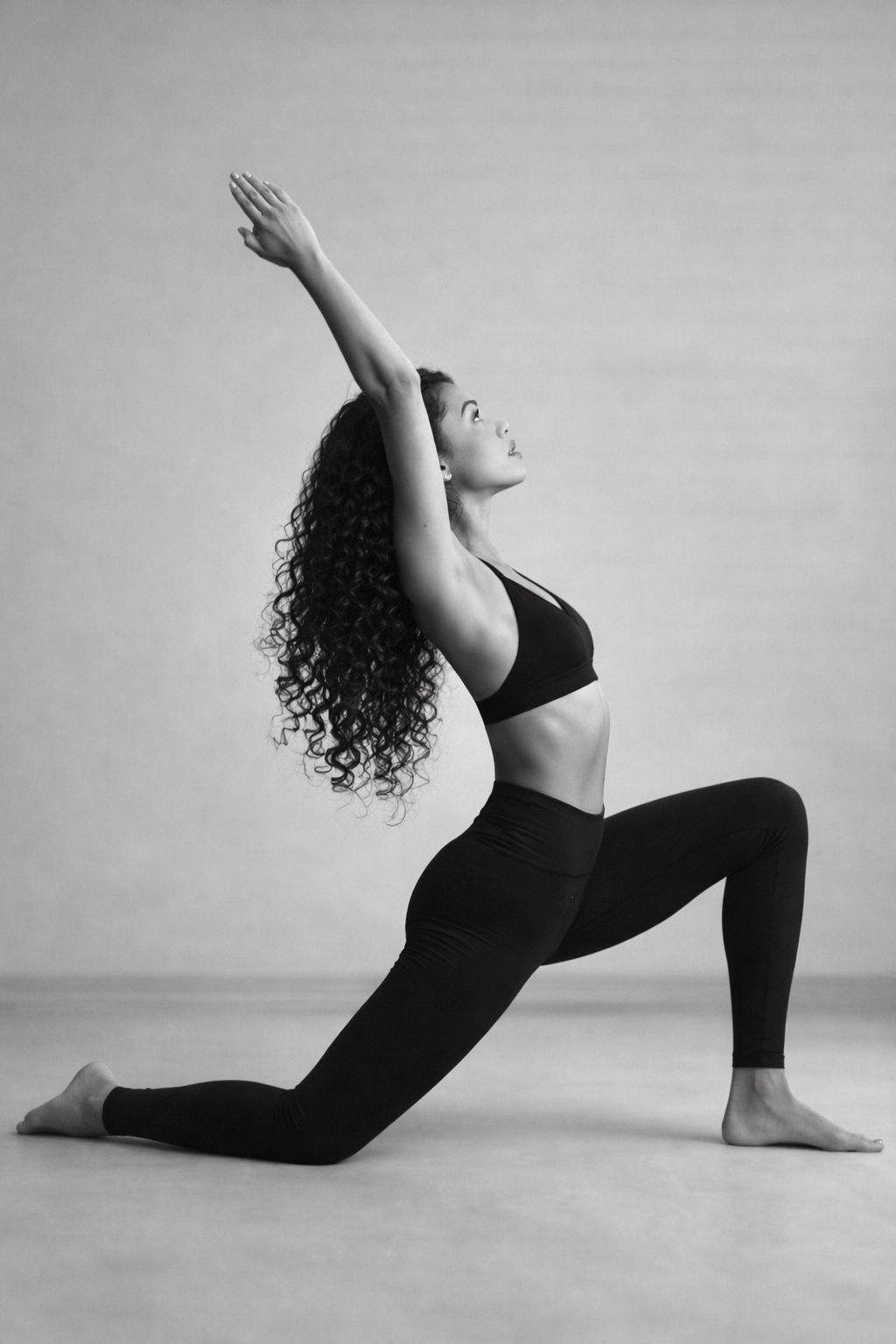 Sophia in a crescent low lunge, arms lifted and arcing back — studio portrait in black and white.