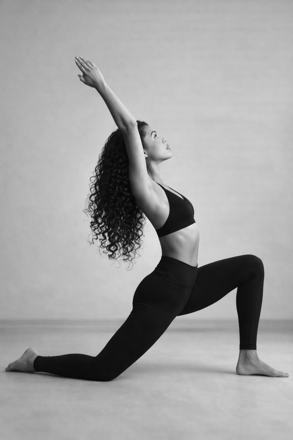 Sophia in a crescent low lunge, arms lifted and arcing back — studio portrait in black and white.